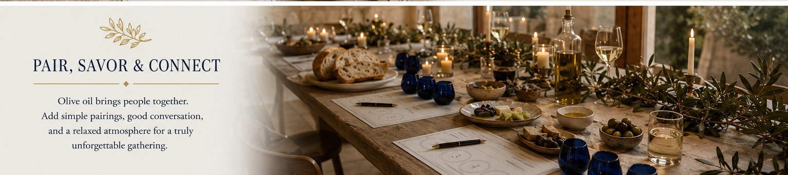 Pair, Savor and Connect — a long candlelit wooden table set for an olive oil tasting, with multiple guests' tasting cards laid out at each place, cobalt blue tasting cups, sliced bread, small dishes of olives, olive branches running down the center, candles, an open oil bottle, and stemware in the background