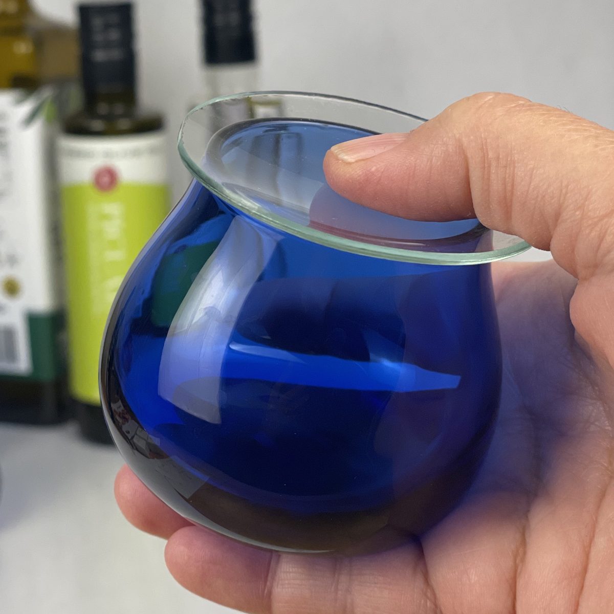 A hand holding a cobalt-blue olive oil tasting glass with a clear glass lid resting on top